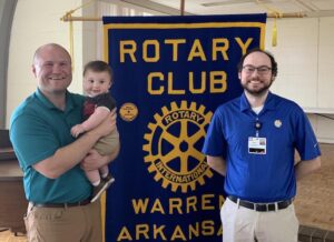 Two men holding a baby in front of a rotary banner.