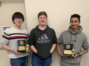 Three young men holding trophies in front of a wall.