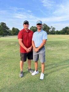 Two men standing on a golf course with trees in the background.