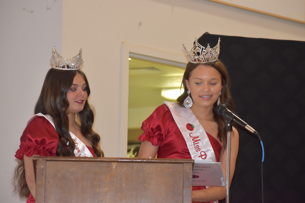Two women in red dresses and tiaras are speaking at a podium.