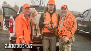 Three men in orange jackets standing next to a dog.