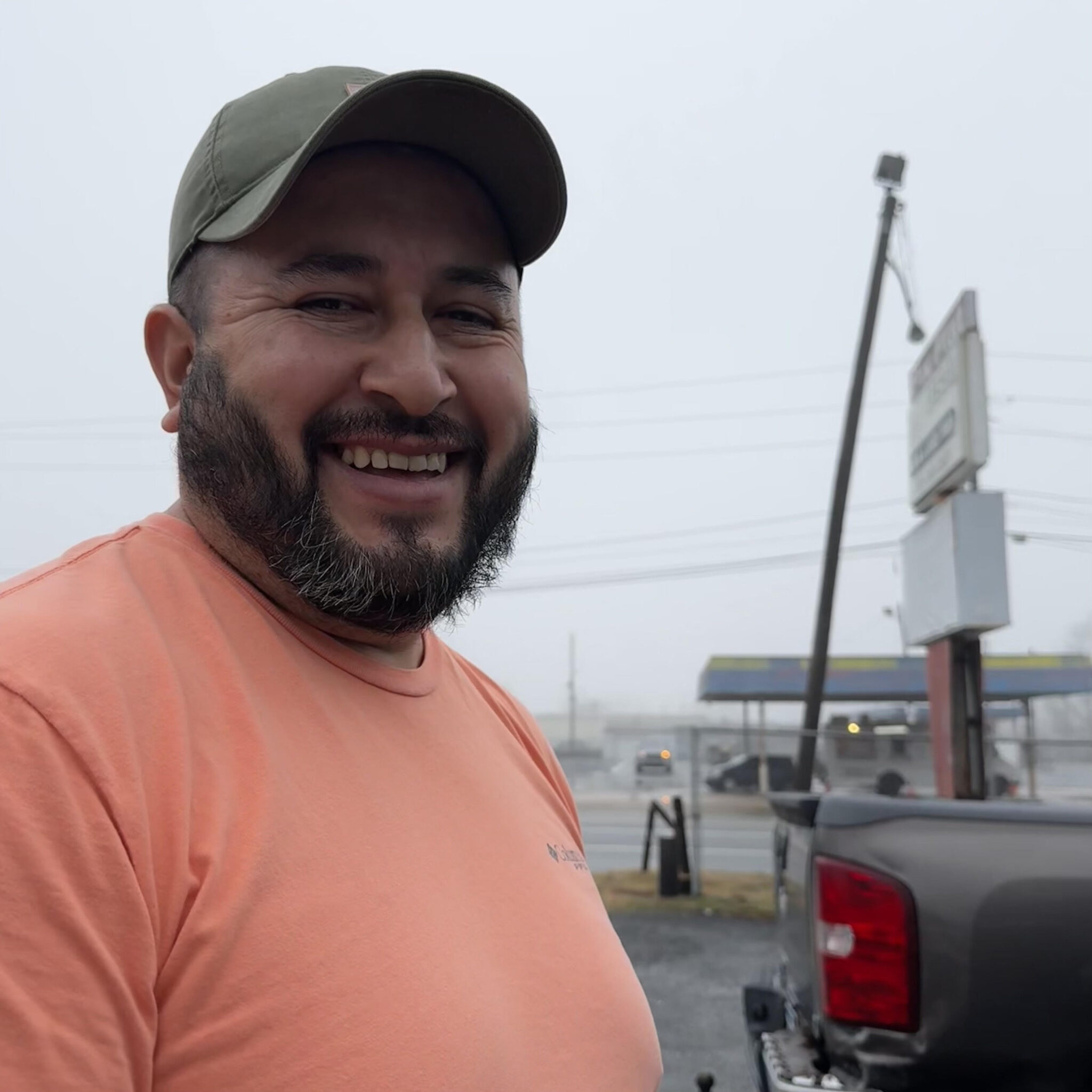 A man in an orange shirt and hat standing next to a truck.